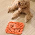 Small dog sitting on a wooden floor next to an orange lick mat with treats on it.